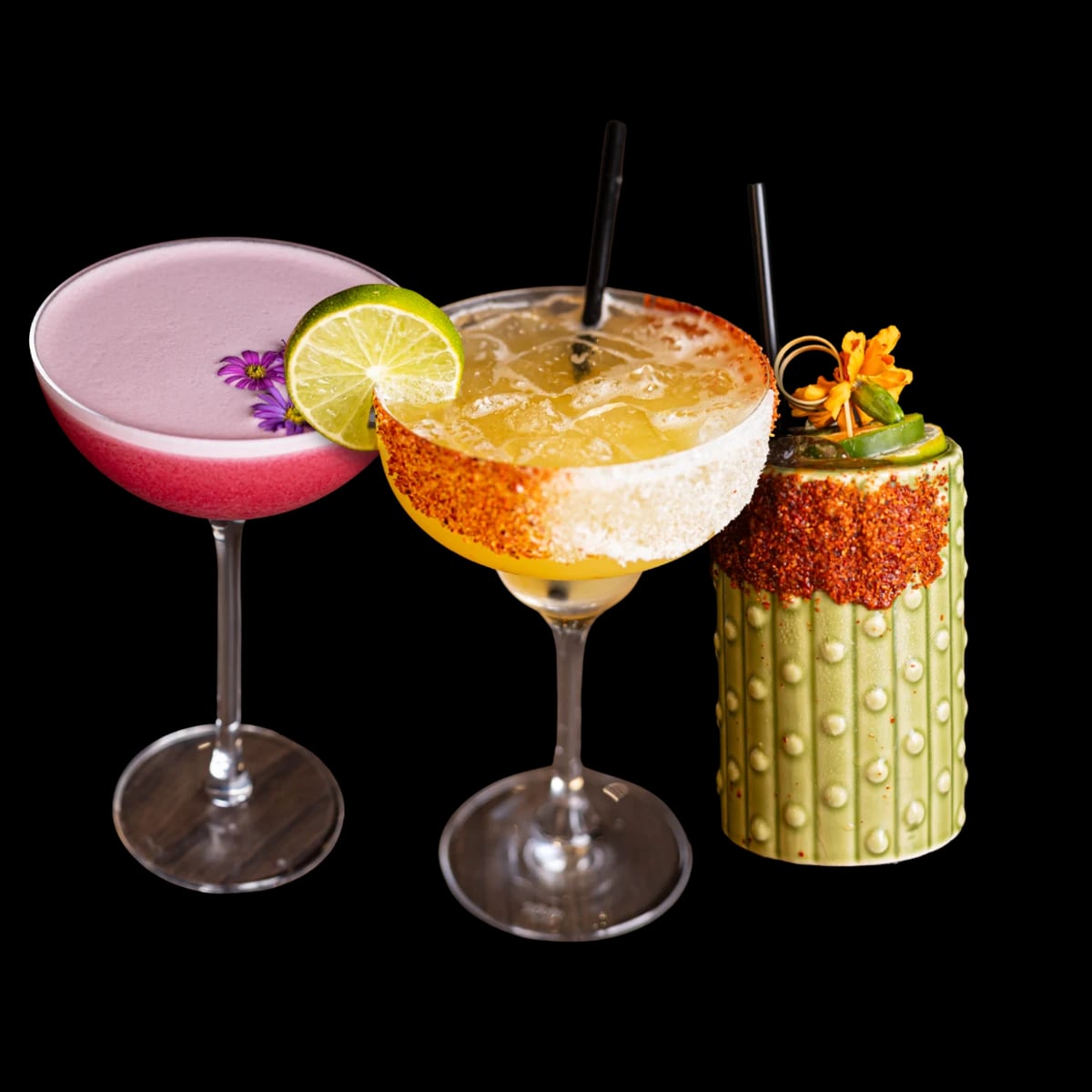 Three cocktails: a pink drink in a coupe with purple flowers, a margarita with a chile and salt rim, and a drink in a green cactus-shaped mug